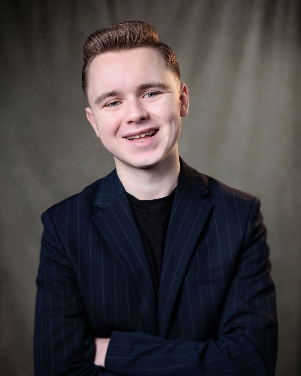Professional headshot of someone wearing a navy pinstriped blazer and black shirt against a gray background.