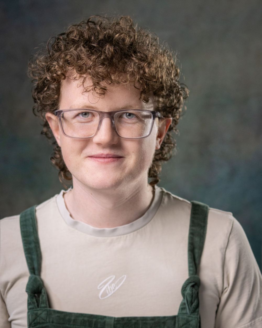 Studio portrait of a person with curly hair wearing glasses and overalls against a dark background.