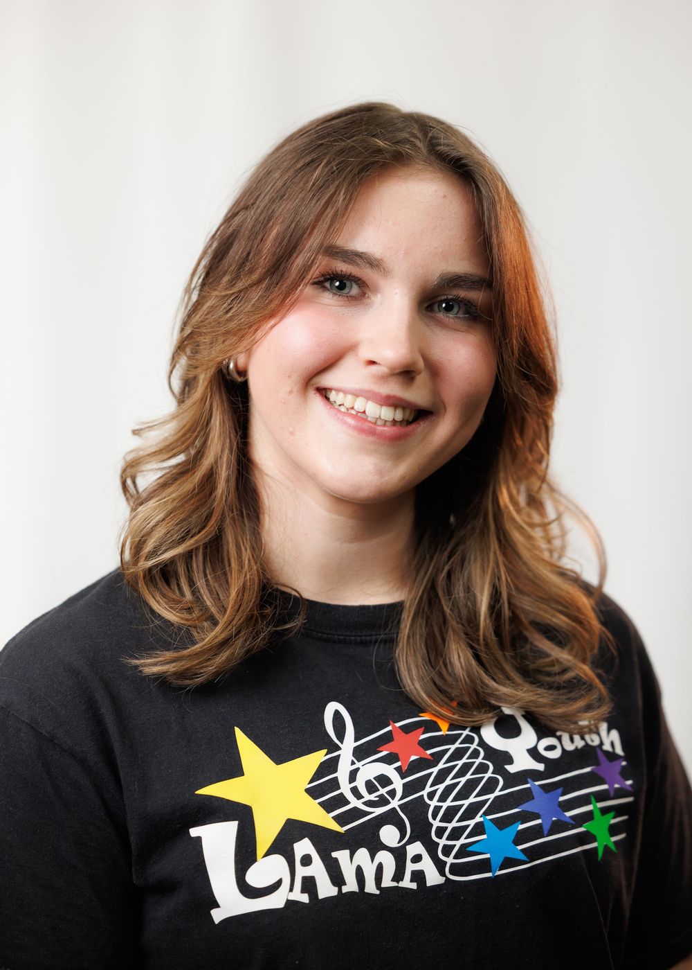 Smiling young woman with wavy brown hair wearing a black LAMA Youth music program t-shirt.