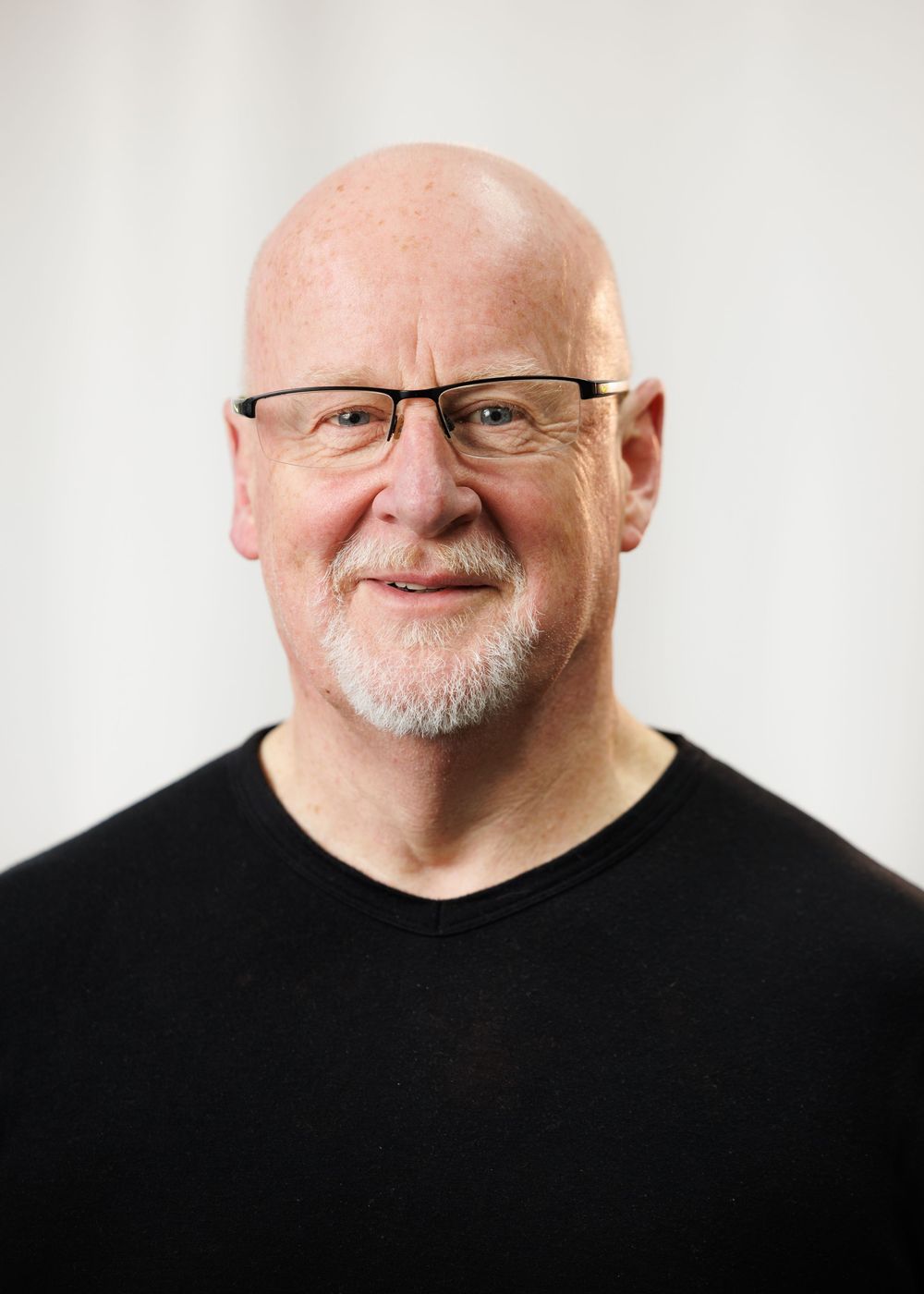 Bald older man with glasses and white goatee smiling, wearing a black V-neck shirt against a white background.