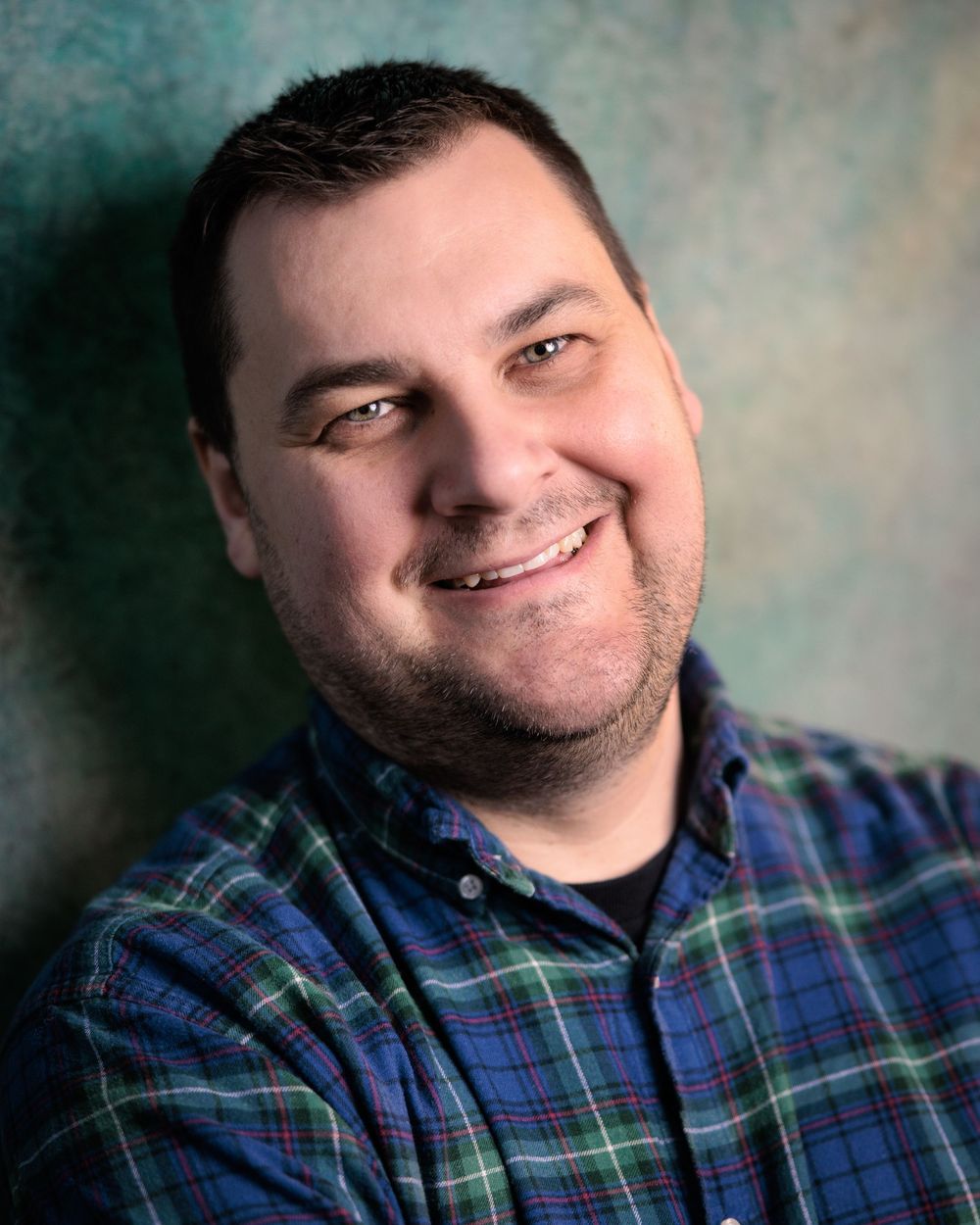 A person wearing a blue plaid button-up shirt smiles warmly against a textured gray background.