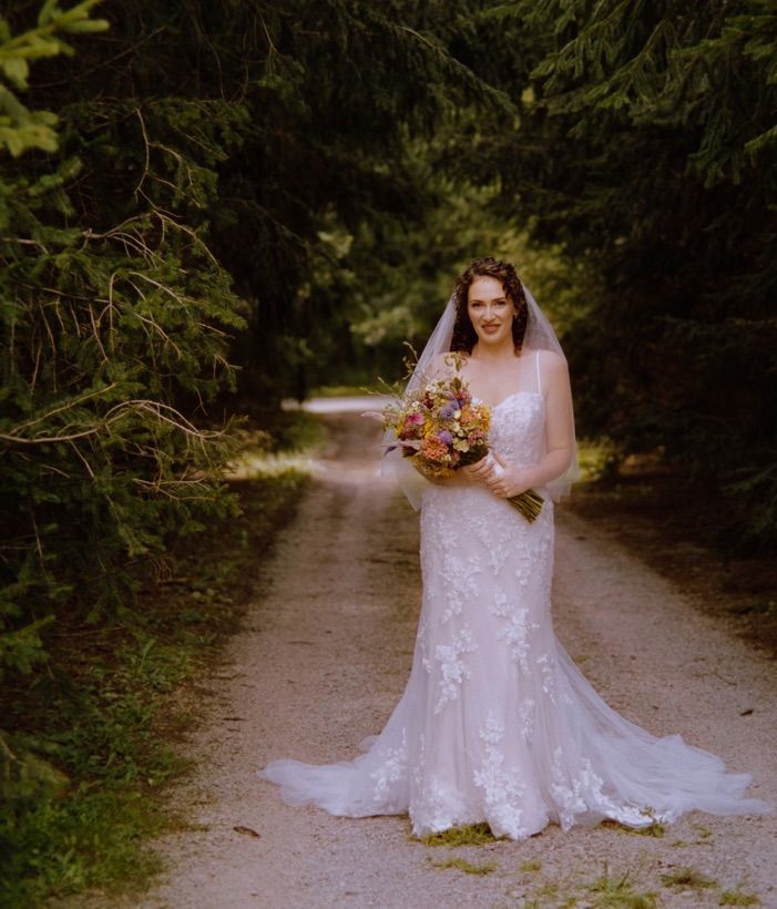 A bride in a white lace gown holds a fall-colored bouquet while posing on a tree-lined path in autumn. sanaview film photos
