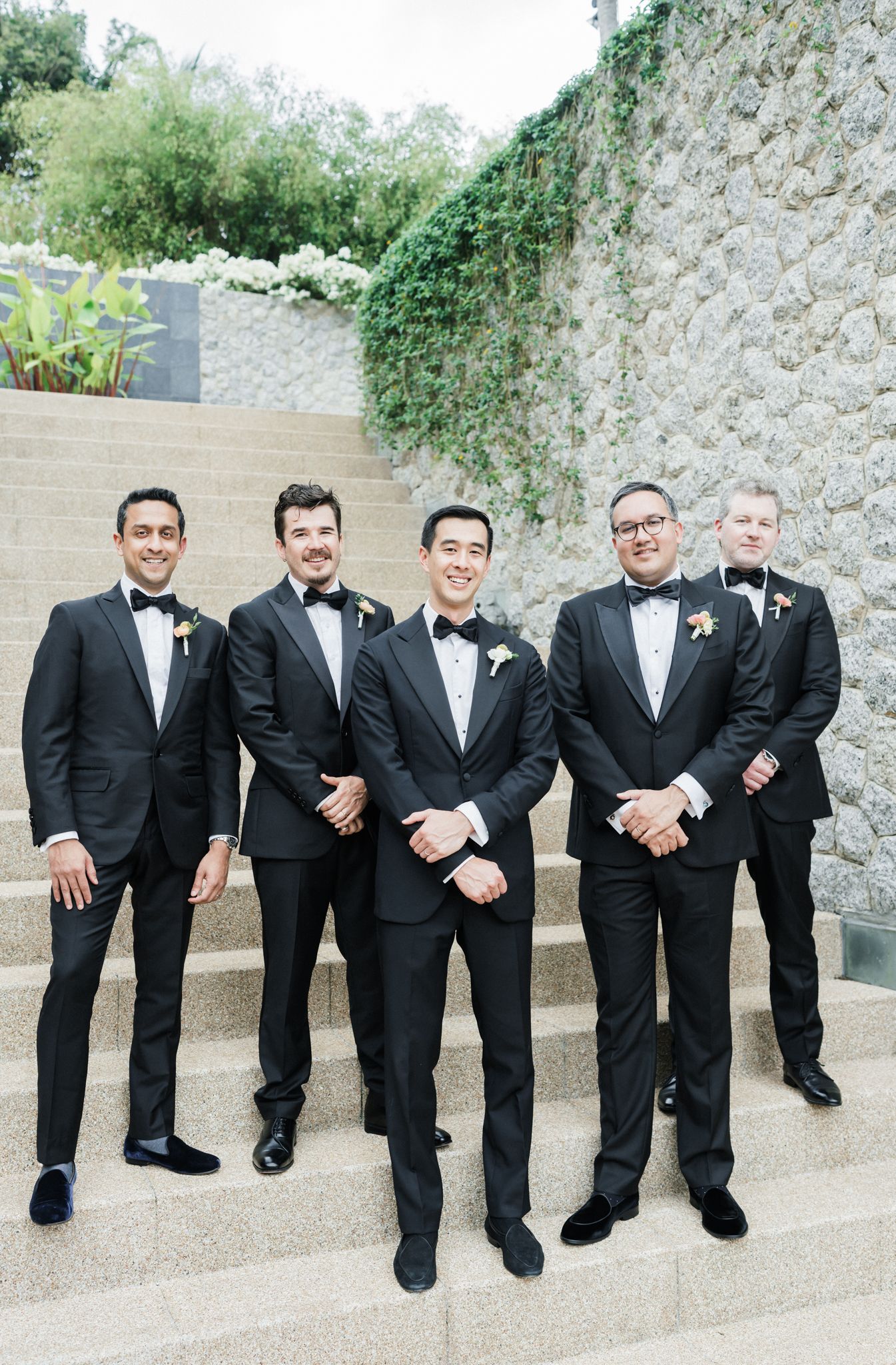 Groomsmen in black tuxedos sitting casually on concrete steps for a group photo.