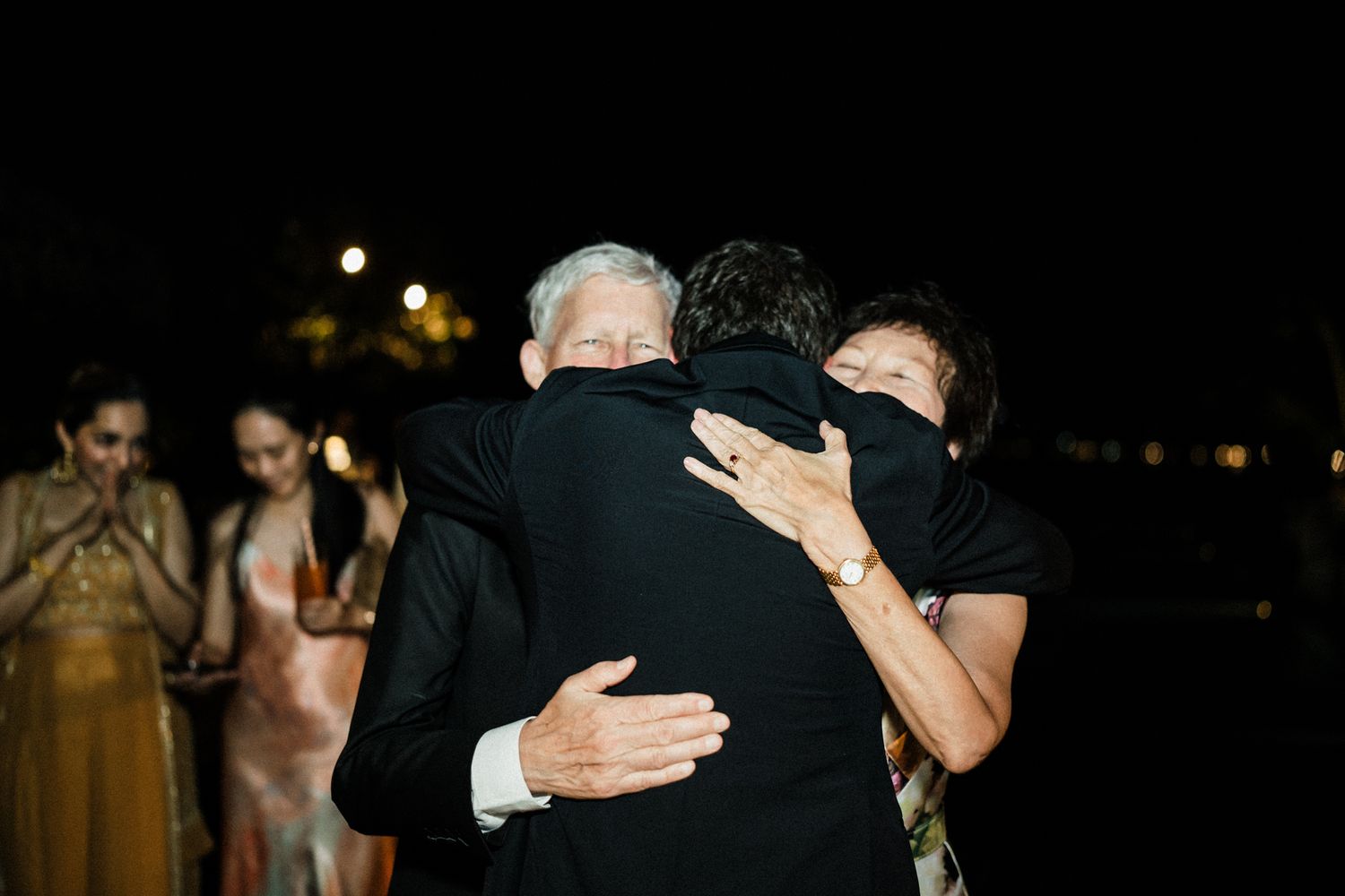A group of people share intimate hugs and embraces in a darkly lit setting during what appears to be a celebration.