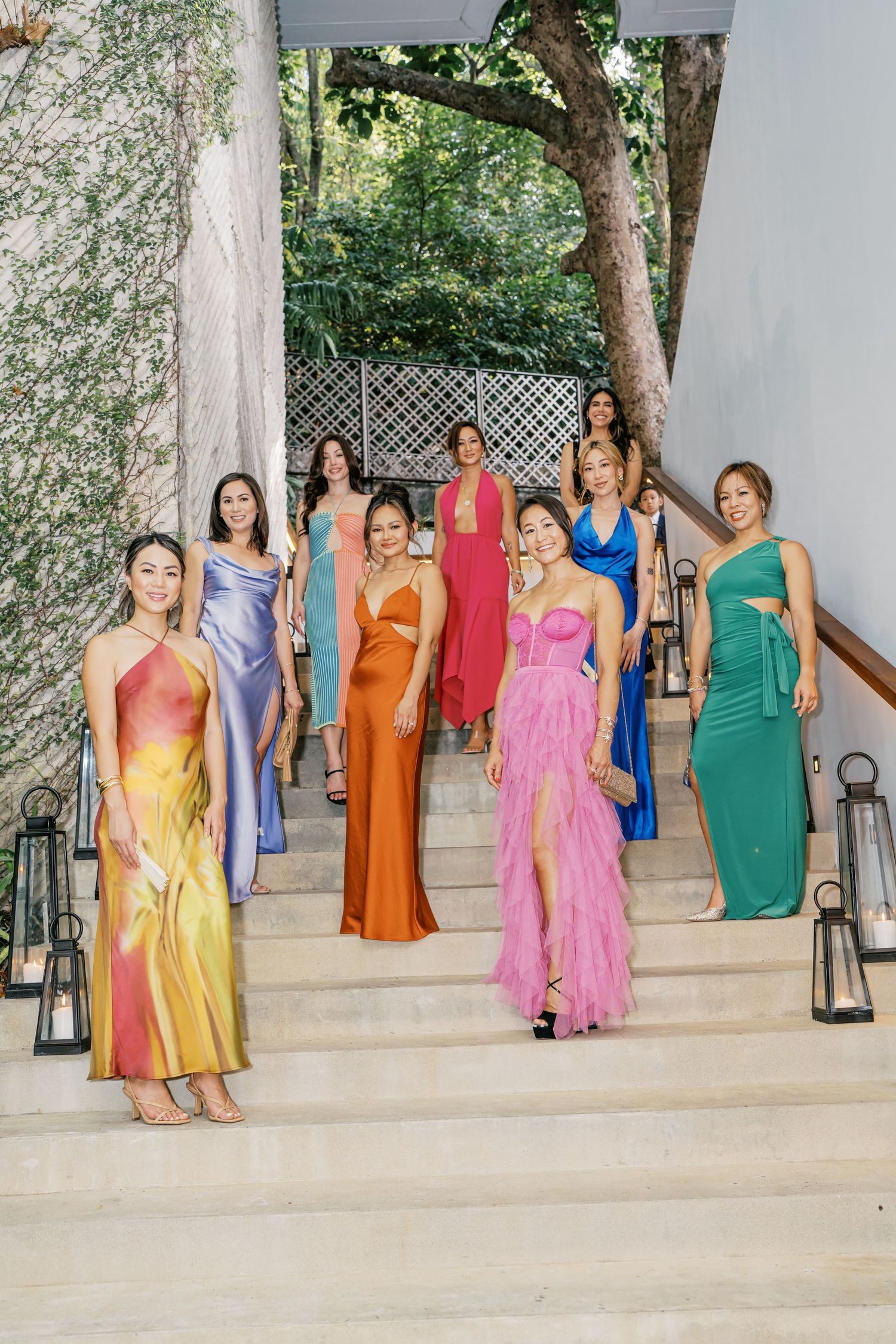 Bridesmaids in colorful satin dresses pose on white stone steps at a garden venue.