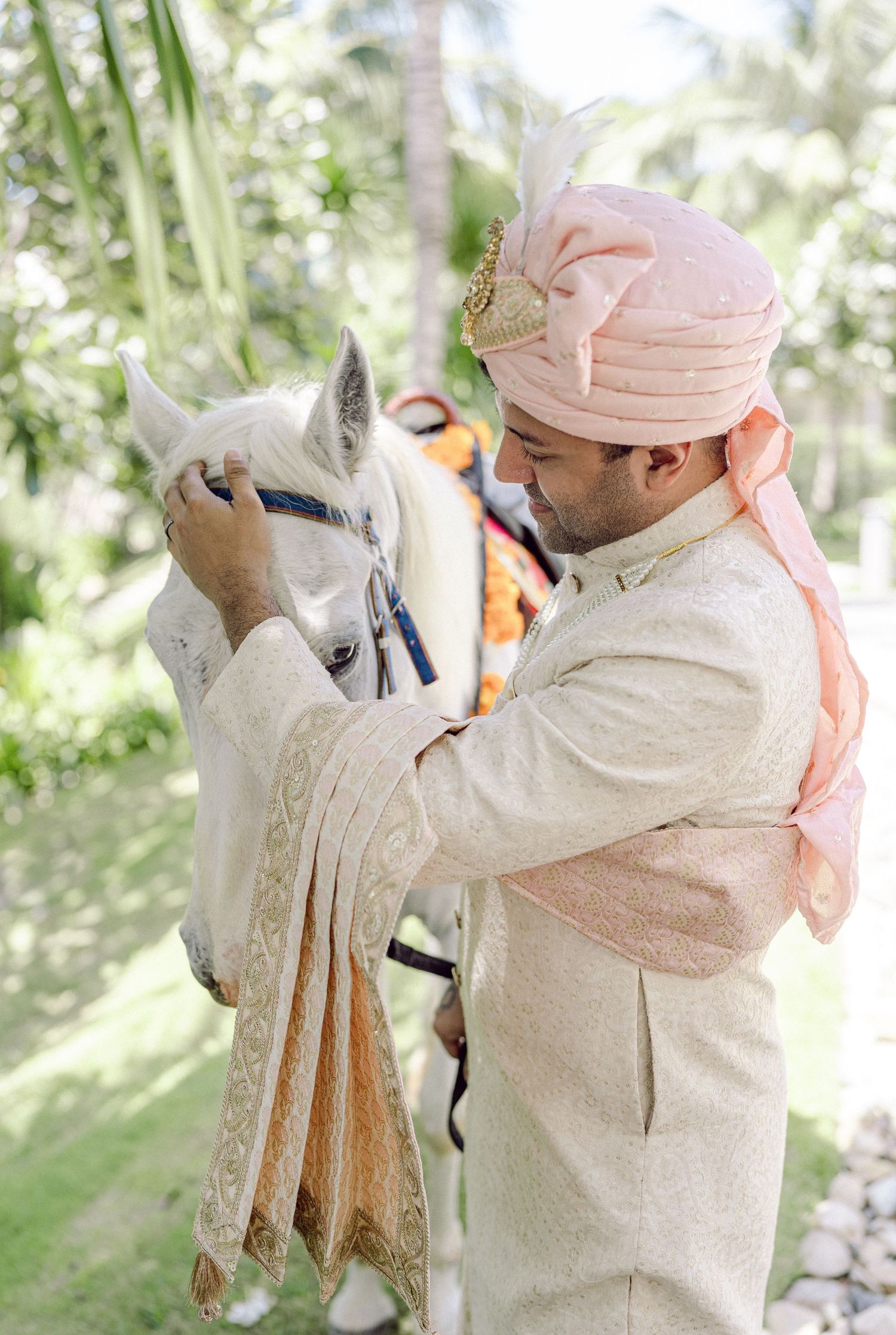 Groom in traditional light-colored attire and pink turban adjusting a white horse's bridle.