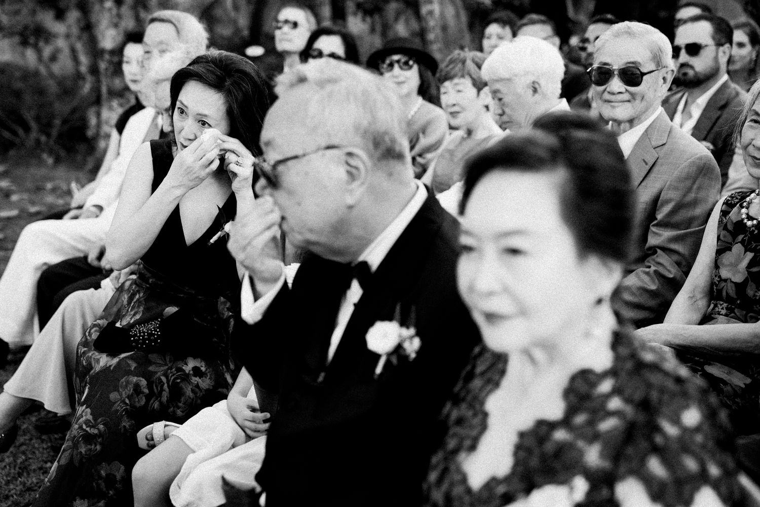Black and white candid shot of wedding guests seated during an outdoor ceremony.