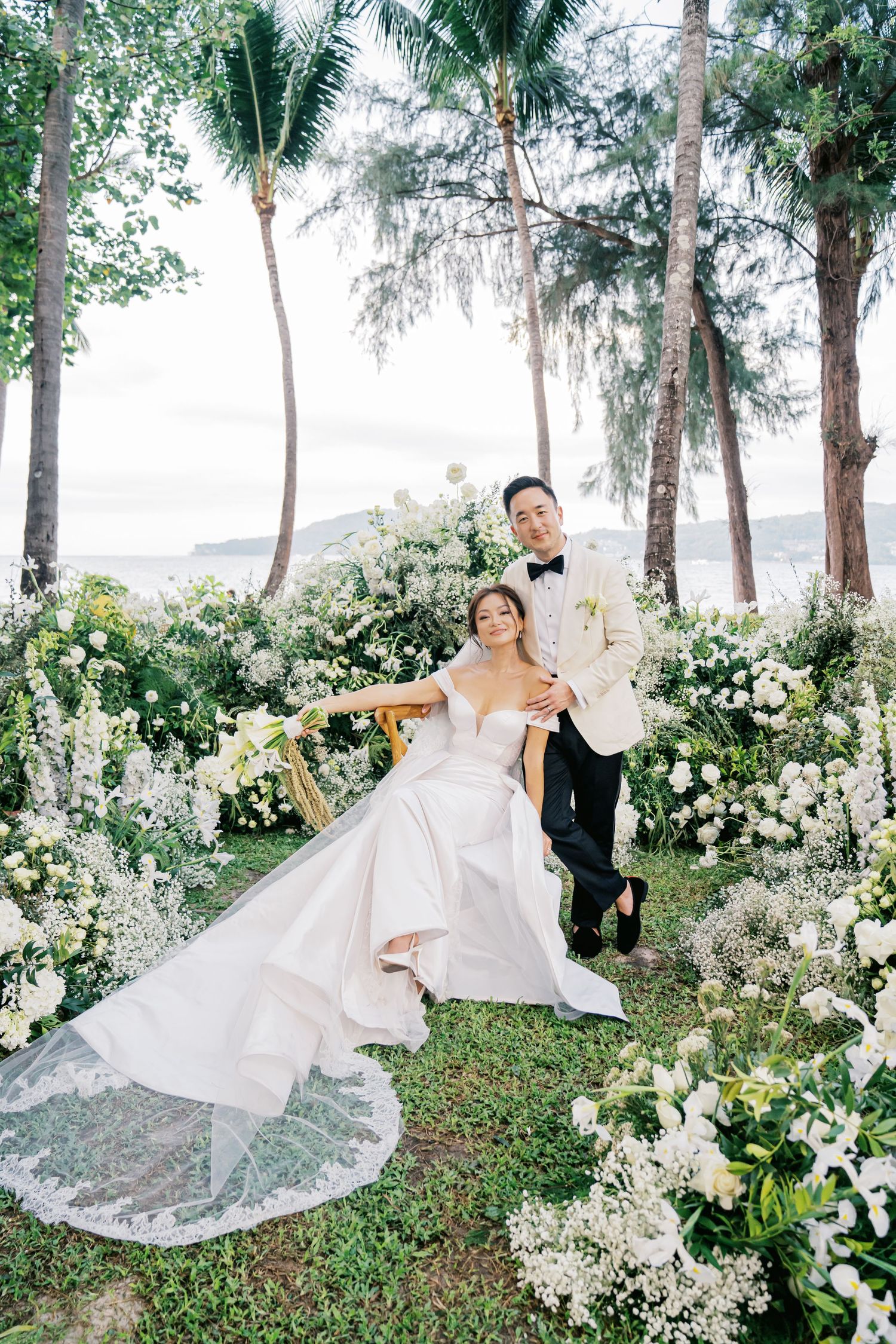 A couple poses among tropical palm trees and white floral arrangements at their beach wedding.
