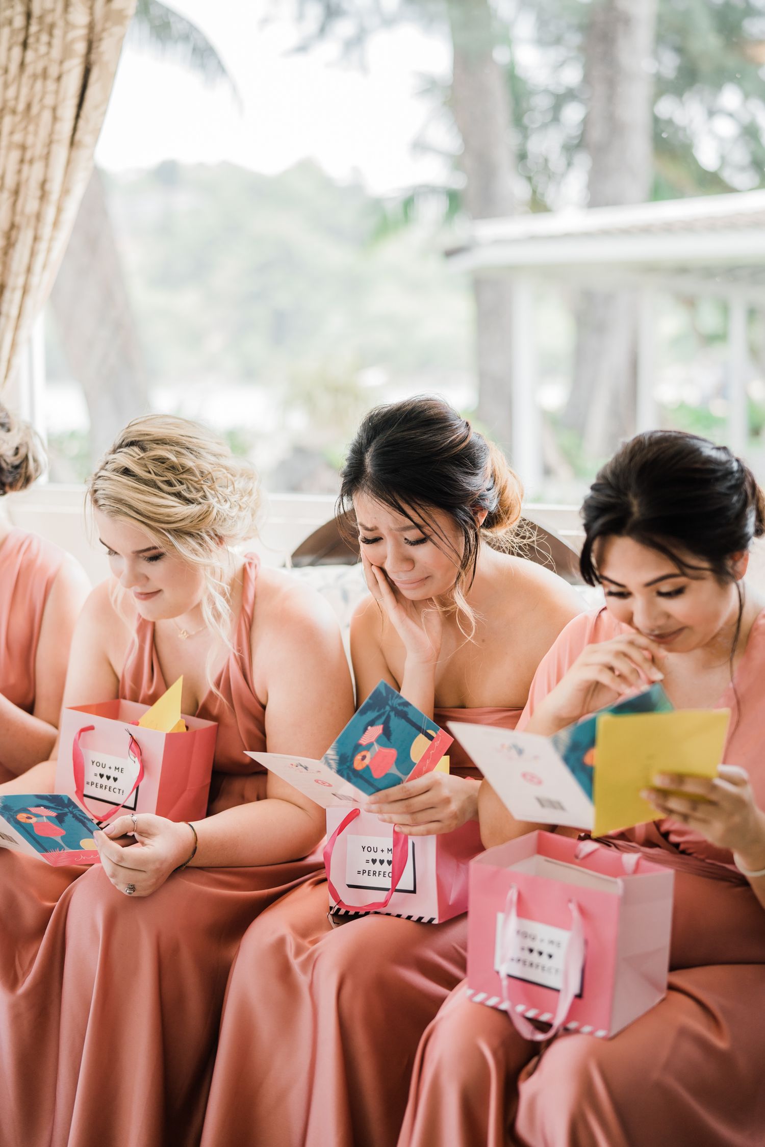Bridesmaids in coral dresses sitting in a row opening gift boxes together.