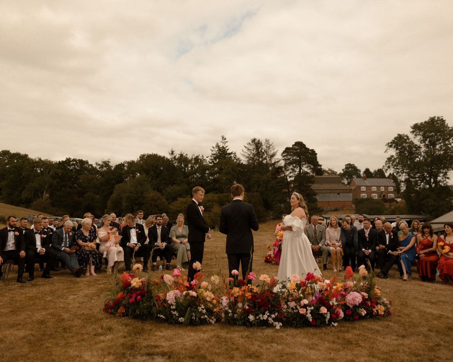 A cheerful wedding party celebrates on the grounds of a historic stone manor house in muted sepia tones.