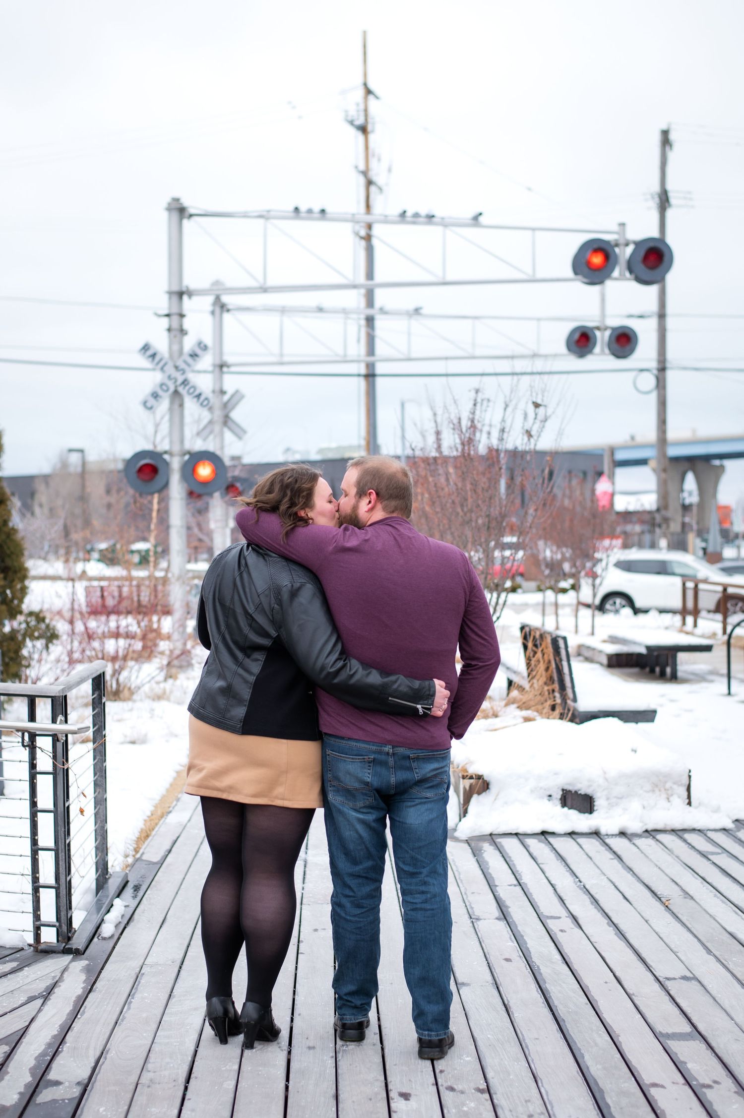 A couple shares a romantic moment while standing on a wooden dock near an industrial bridge structure.