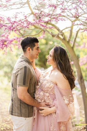 A romantic maternity photoshoot under blooming cherry blossom trees with soft pink petals and golden sunlight.