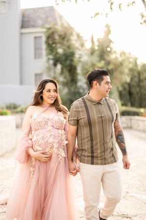 A couple walking outdoors at sunset with the woman wearing a flowing pink tulle gown and cradling her pregnant belly.