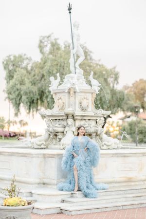 Woman in ethereal blue gown poses by ornate white fountain in dreamy outdoor setting.
