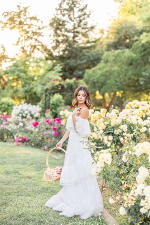 Woman in flowing white dress twirls through garden with blooming roses at sunset.