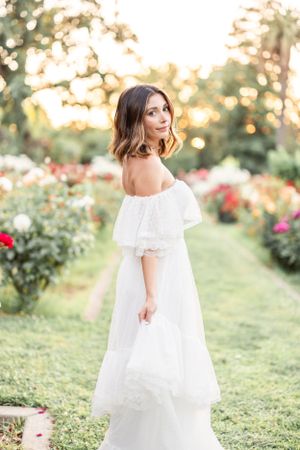 Woman in white off-shoulder dress poses in rose garden during golden hour photoshoot.