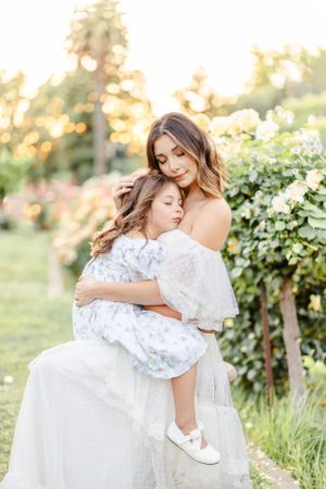 Mother and daughter share tender embrace in white dresses among garden flowers.