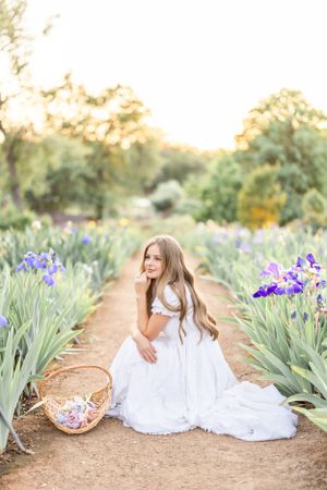 White-dressed figures resting in a garden path surrounded by blooming purple iris flowers at sunset.
