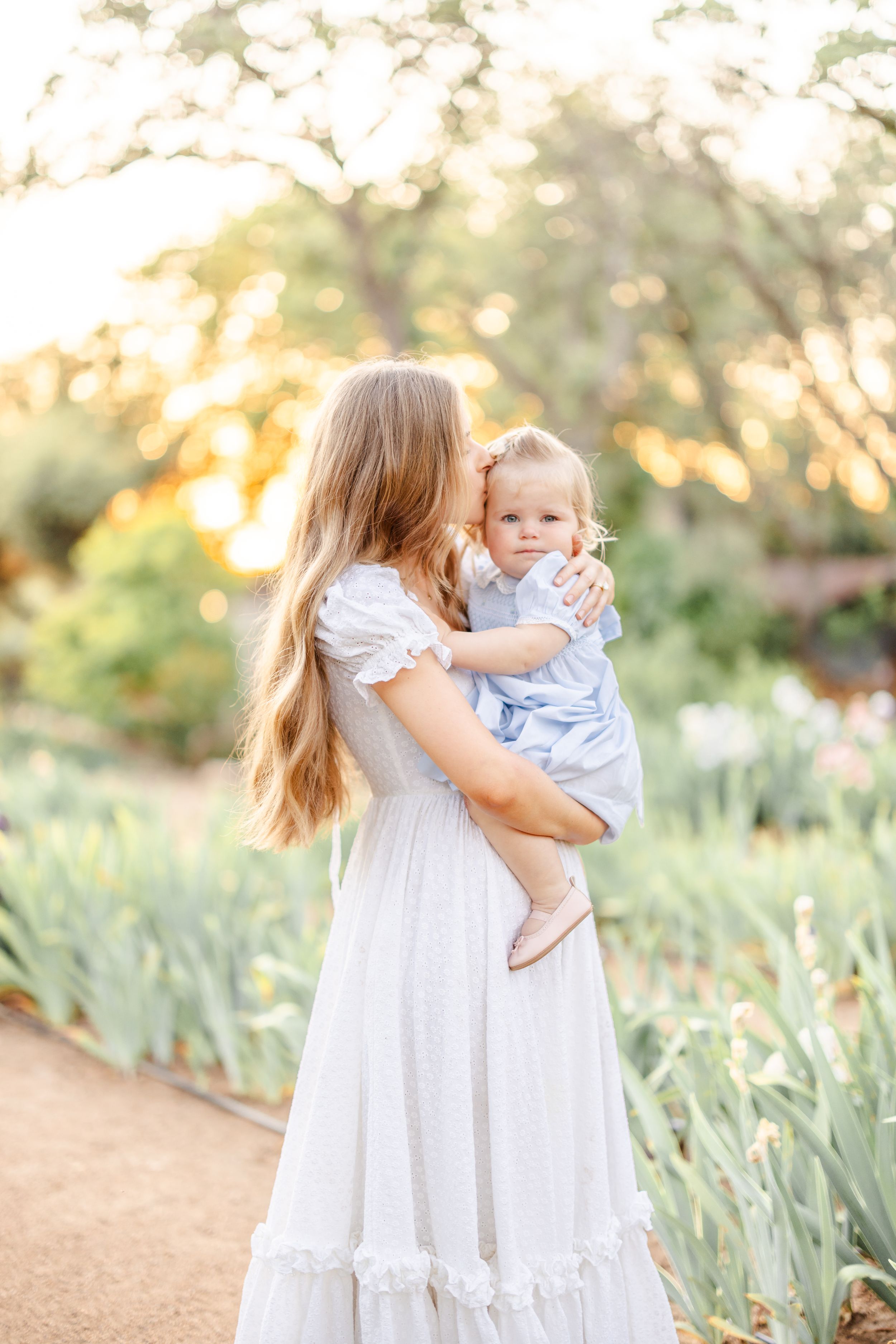 Two people in white dresses share tender moments among blooming flowers during a golden hour photoshoot.