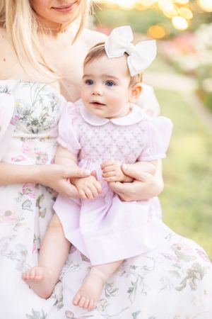 Baby wearing pink and white outfit with bow sits on blanket during outdoor photoshoot.