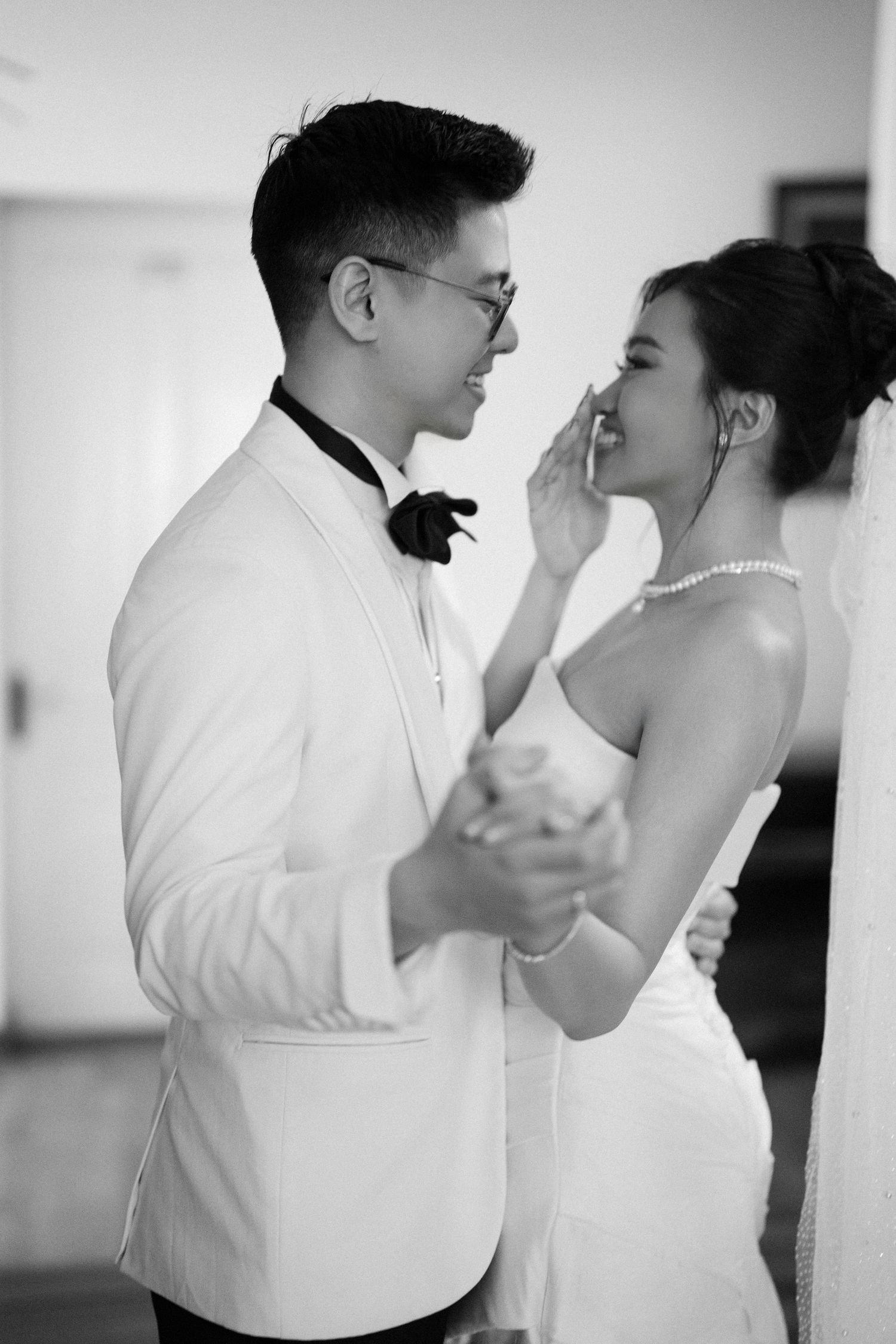 Groom in white tuxedo tenderly touches brides face as they share an intimate black and white moment.