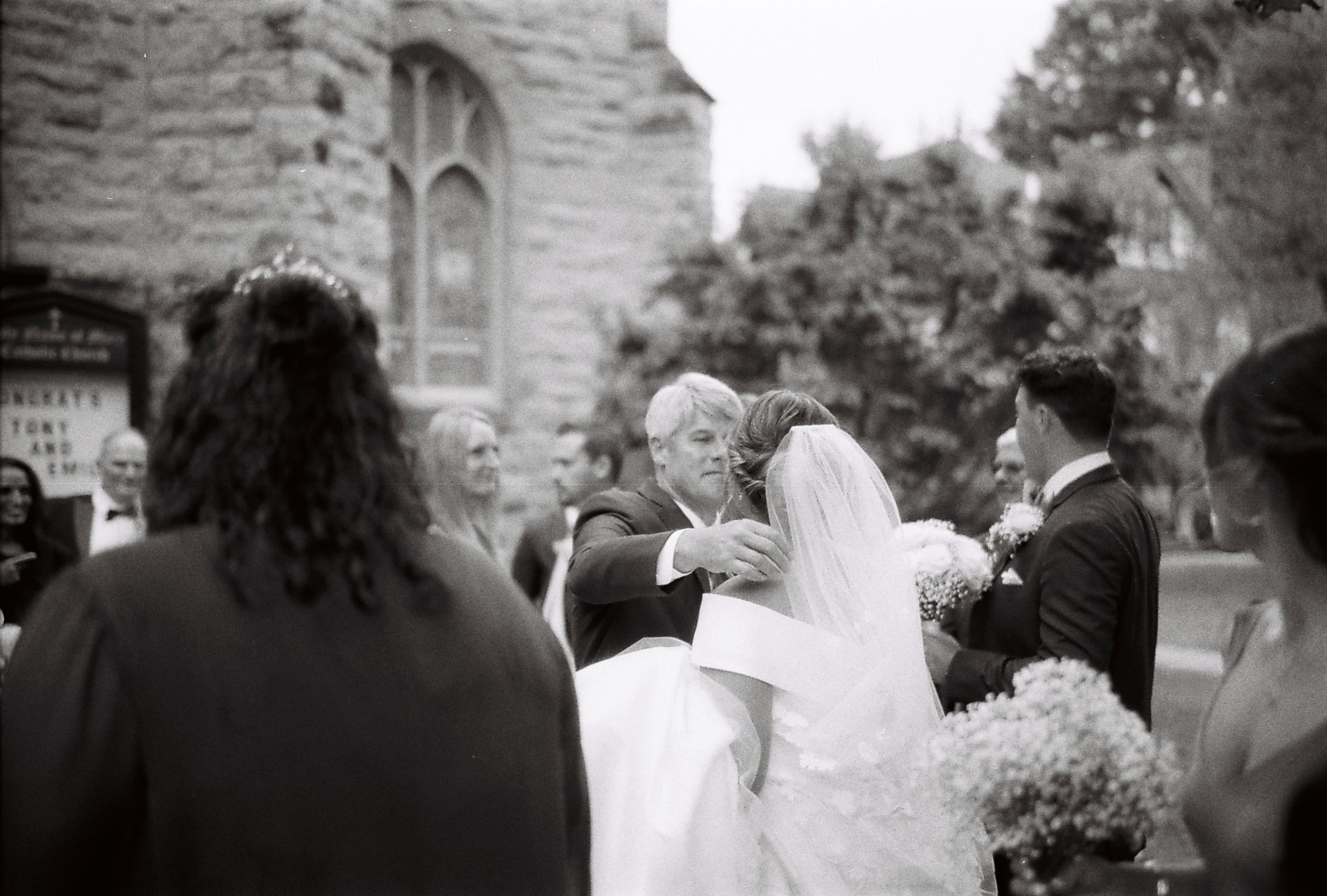 Wedding party and guests mingling outside a stone church building on a sunny day.