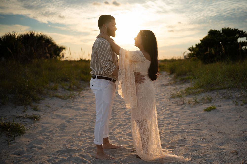 Two romantic figures in white attire walk along a sandy beach path at sunset.