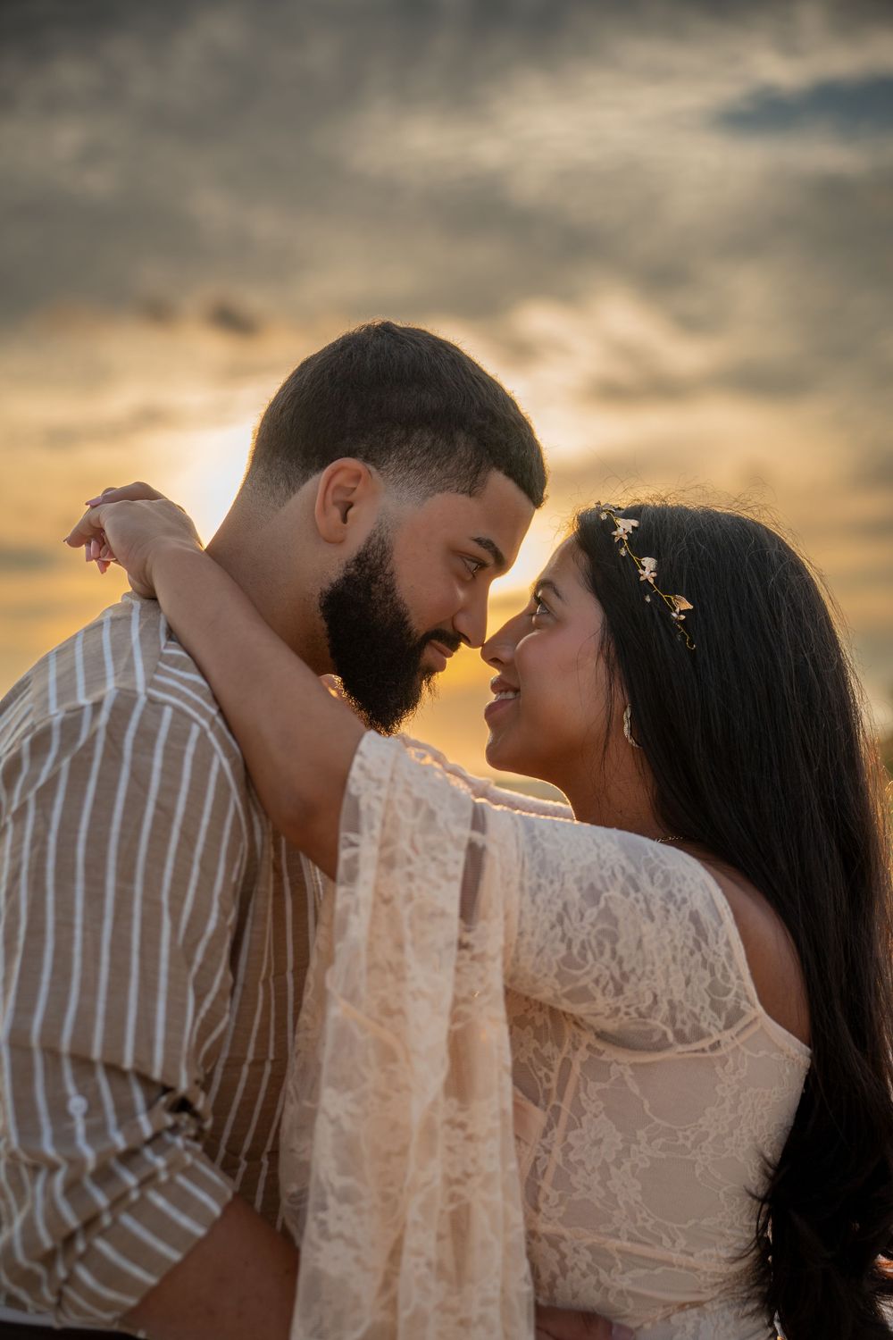 Romantic couple embraces against a dramatic sunset sky with warm golden lighting and soft clouds in background.