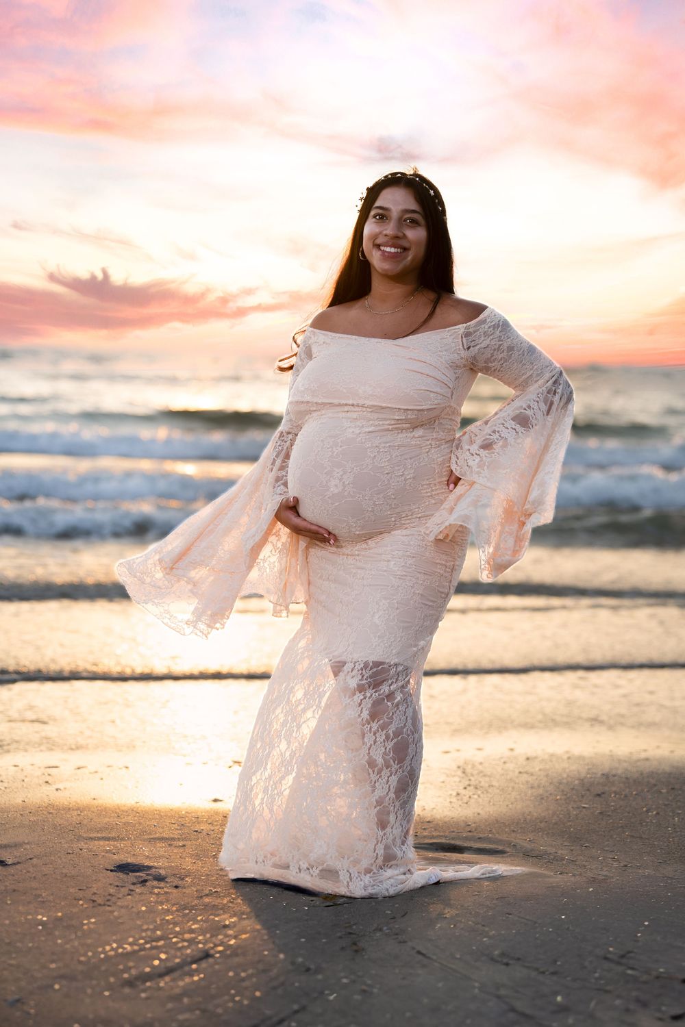 Off-shoulder lace gown accentuates an elegant maternity shoot along the shoreline at sunset with gentle waves rolling in.