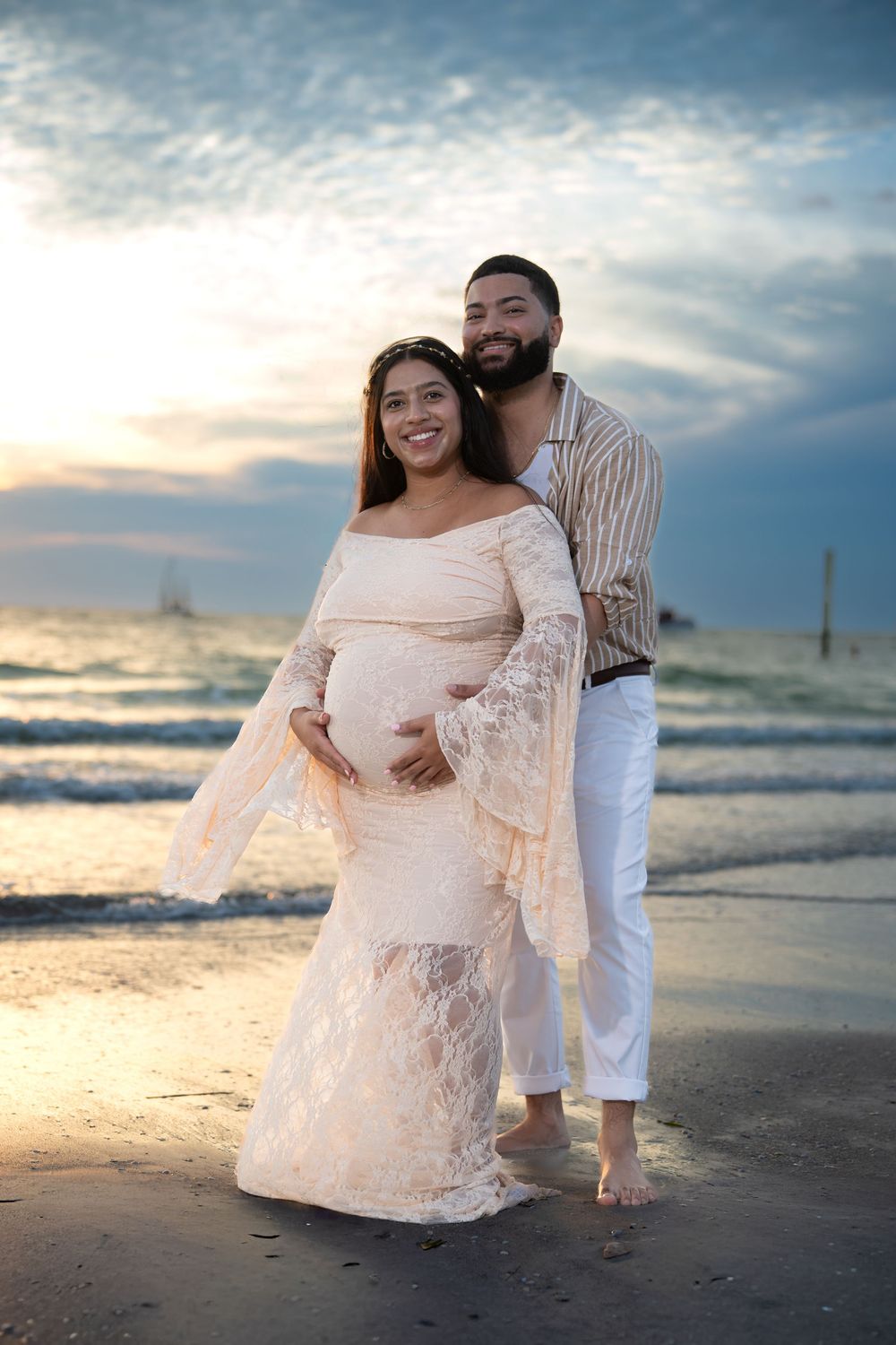 A couple shares intimate moments on a sandy beach during sunset with waves and cloudy sky in the background.