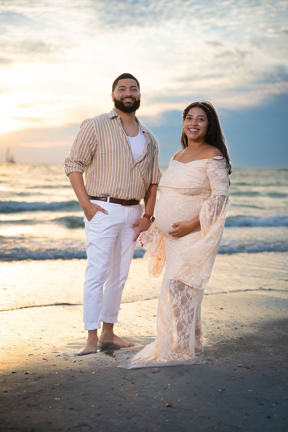A couple poses together on a beach at sunset, with waves behind them and the woman wearing a flowing lace dress.