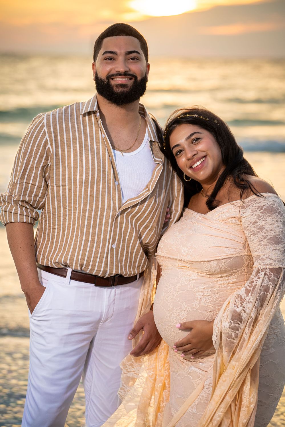 A couple poses together on the beach at sunset wearing coordinated beige and white outfits.