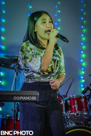Female singer in a patterned green blouse holding a microphone and performing live in front of a red drum set.