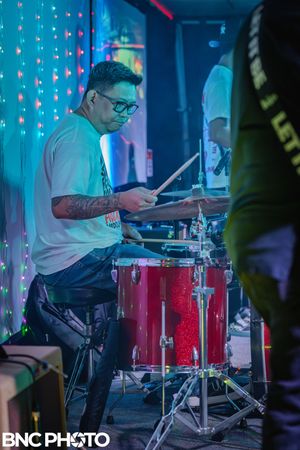 Male drummer playing a red drum kit on stage under colorful lights during a live band performance.