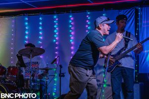Male vocalist in a cap singing into a microphone while band members play drums and bass at a live music show.