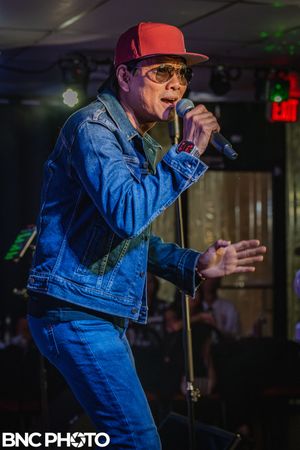 Male vocalist wearing a denim jacket and cap singing on stage during a live music performance at a local venue
