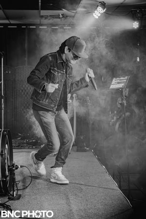 Black and white photo of a male performer in denim jacket and cap singing on stage with dramatic lighting.