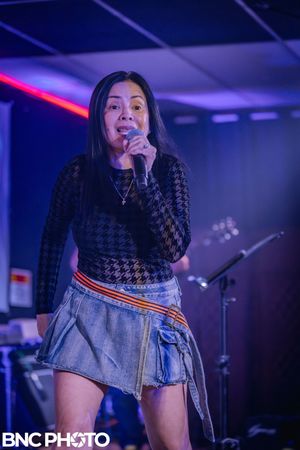 Female singer in a black top and denim skirt performing live on stage with colorful lighting at a concert.