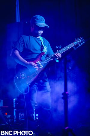 Guitarist wearing a cap playing electric guitar under dramatic blue lighting during a live concert.