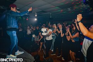 Male performer in denim jacket and cap engaging with fans at a lively indoor music show.