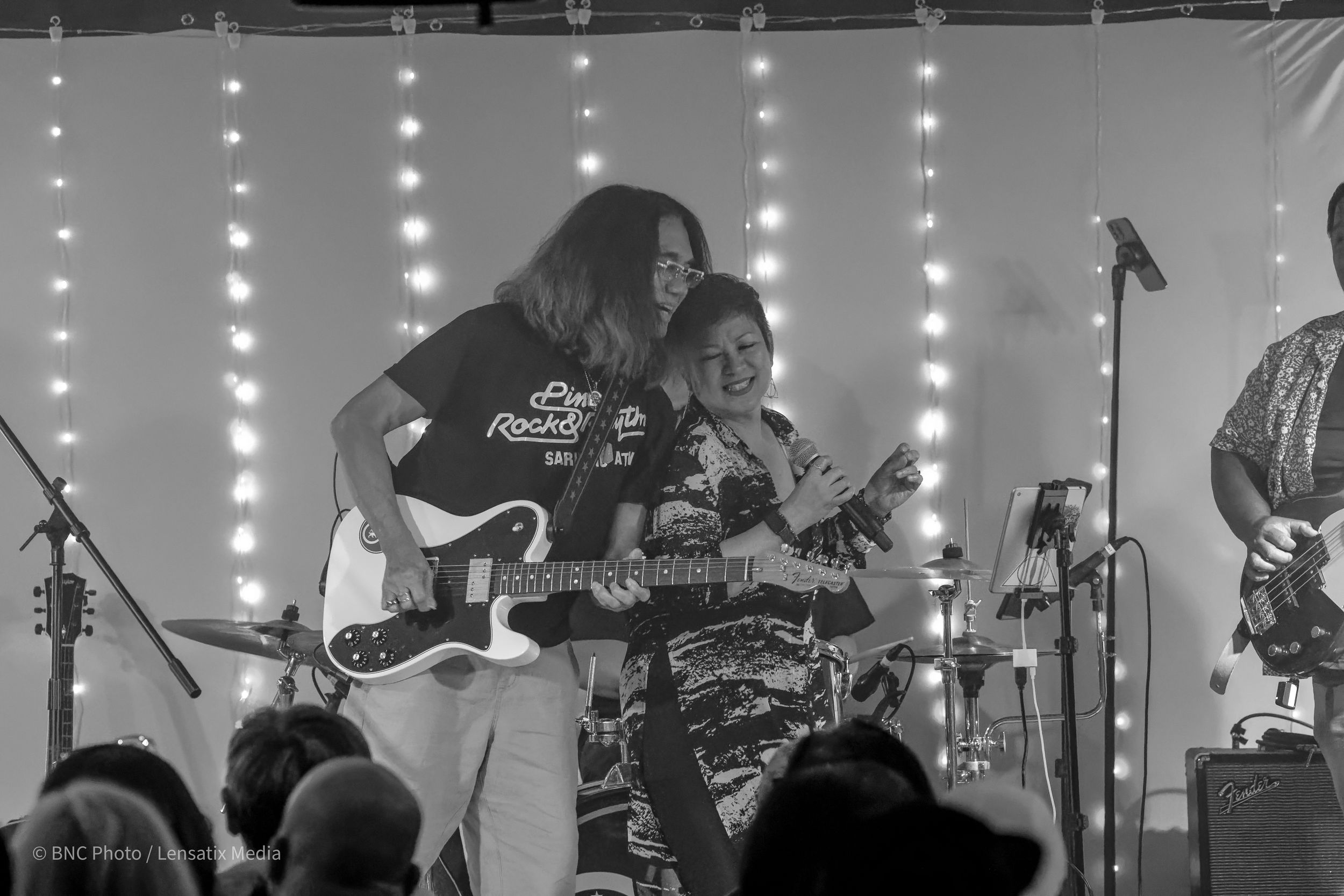 Black and white photo of a guitarist and female singer performing together under string lights at an intimate live music show.