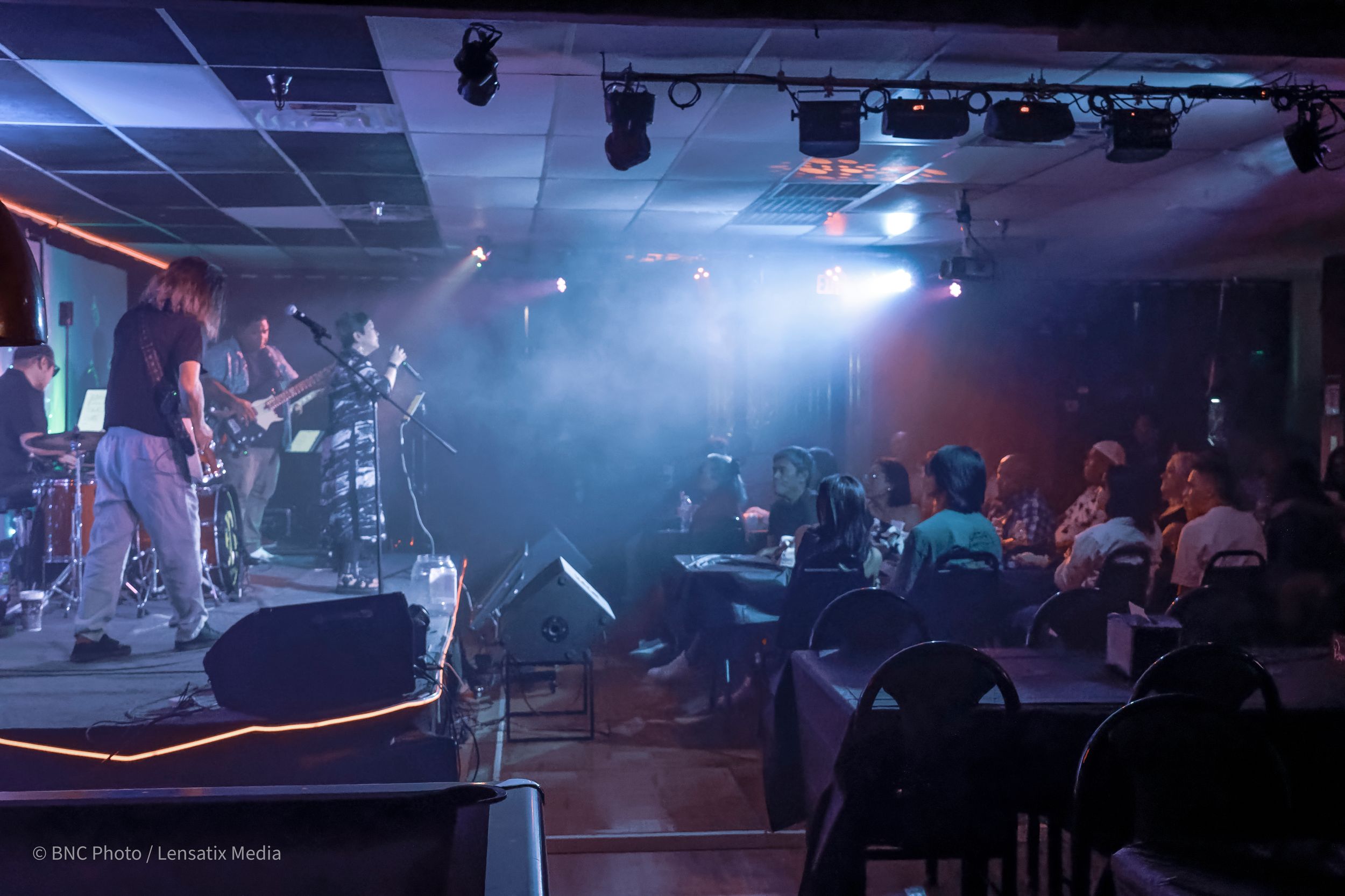 Wide view of a live band performing on stage with colorful lights and fog effects in front of a seated audience at a San Antonio concert.