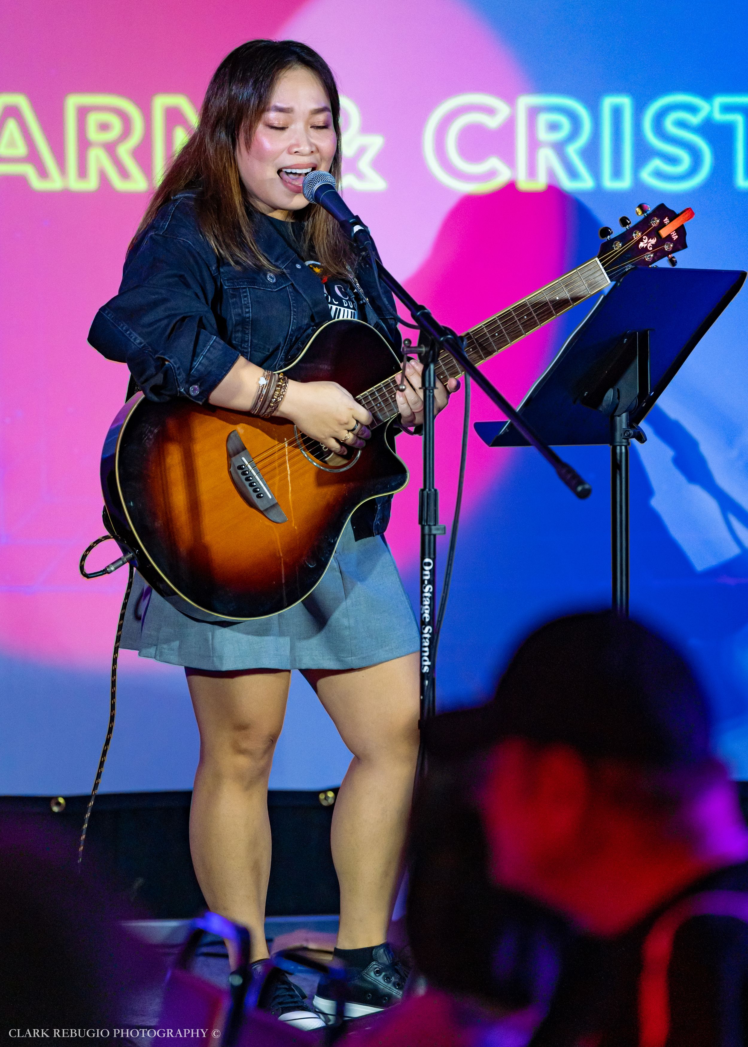 Female musician playing acoustic guitar and singing into a microphone at a live concert with vibrant stage backdrop.