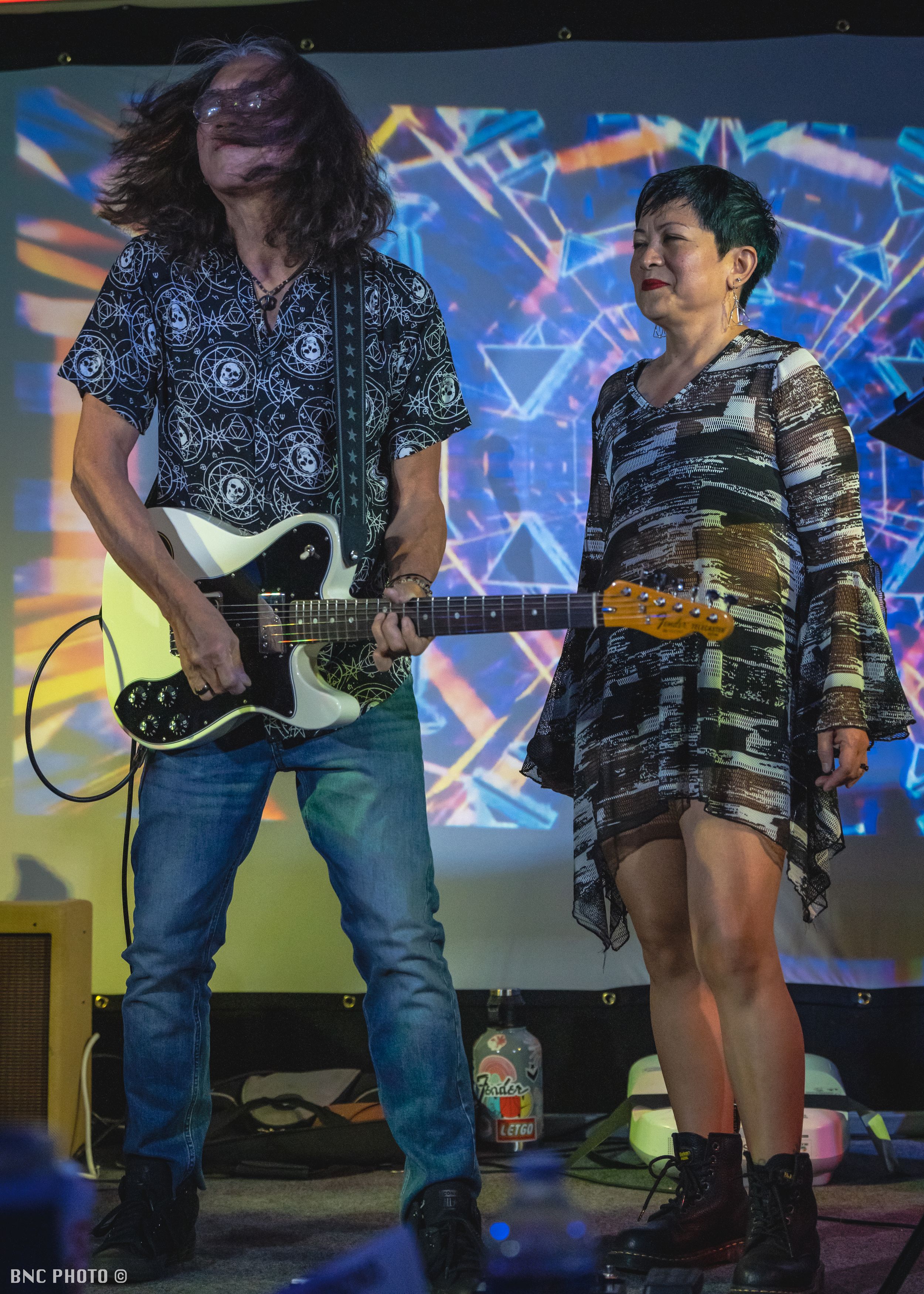 Electric guitarist with long hair performing on stage next to a female singer under vibrant background lights at a live show.