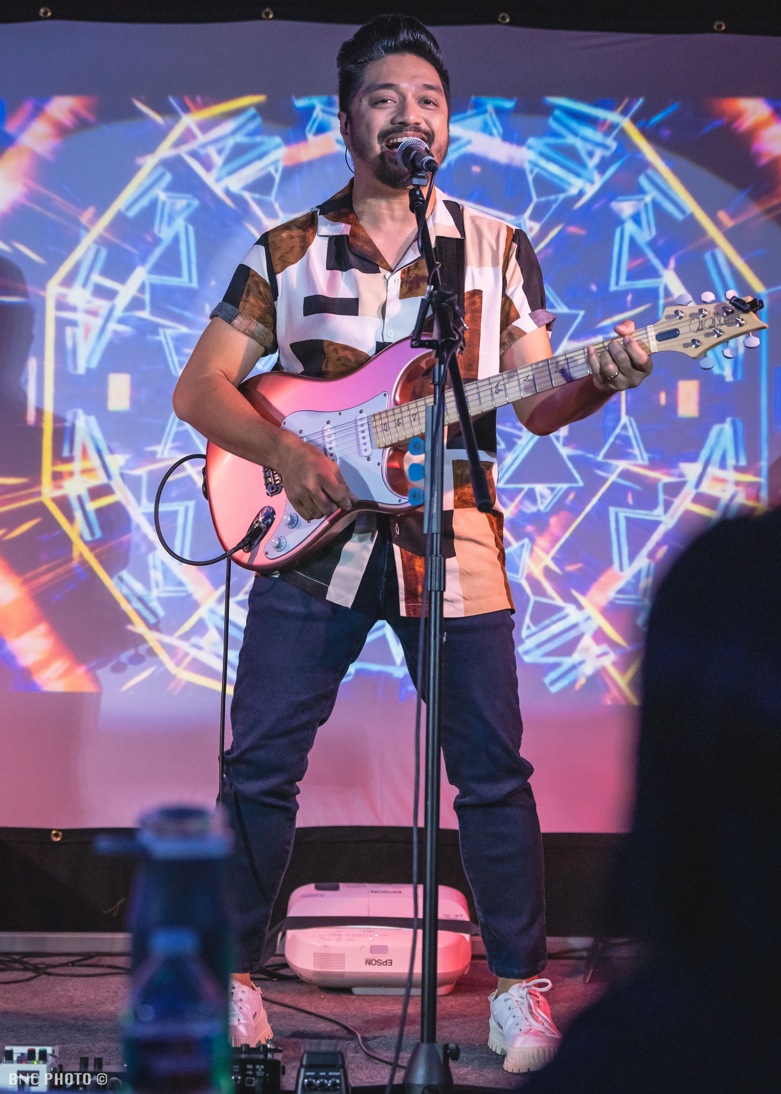Male vocalist playing a pink electric guitar and singing on stage in front of a colorful digital backdrop at a live music performance.