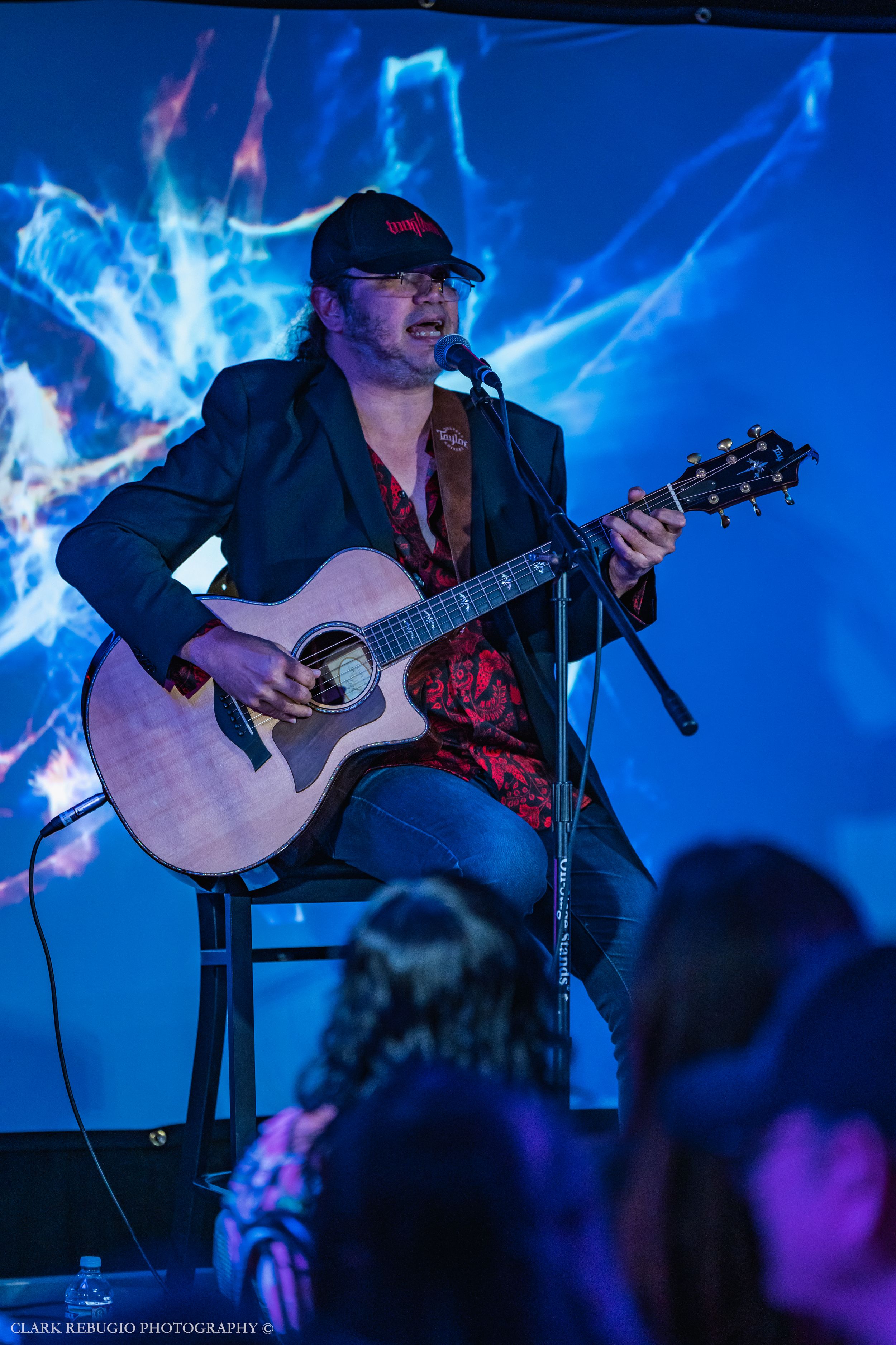 Male musician wearing a cap playing acoustic guitar and singing on stage under dramatic blue lighting at a live concert.