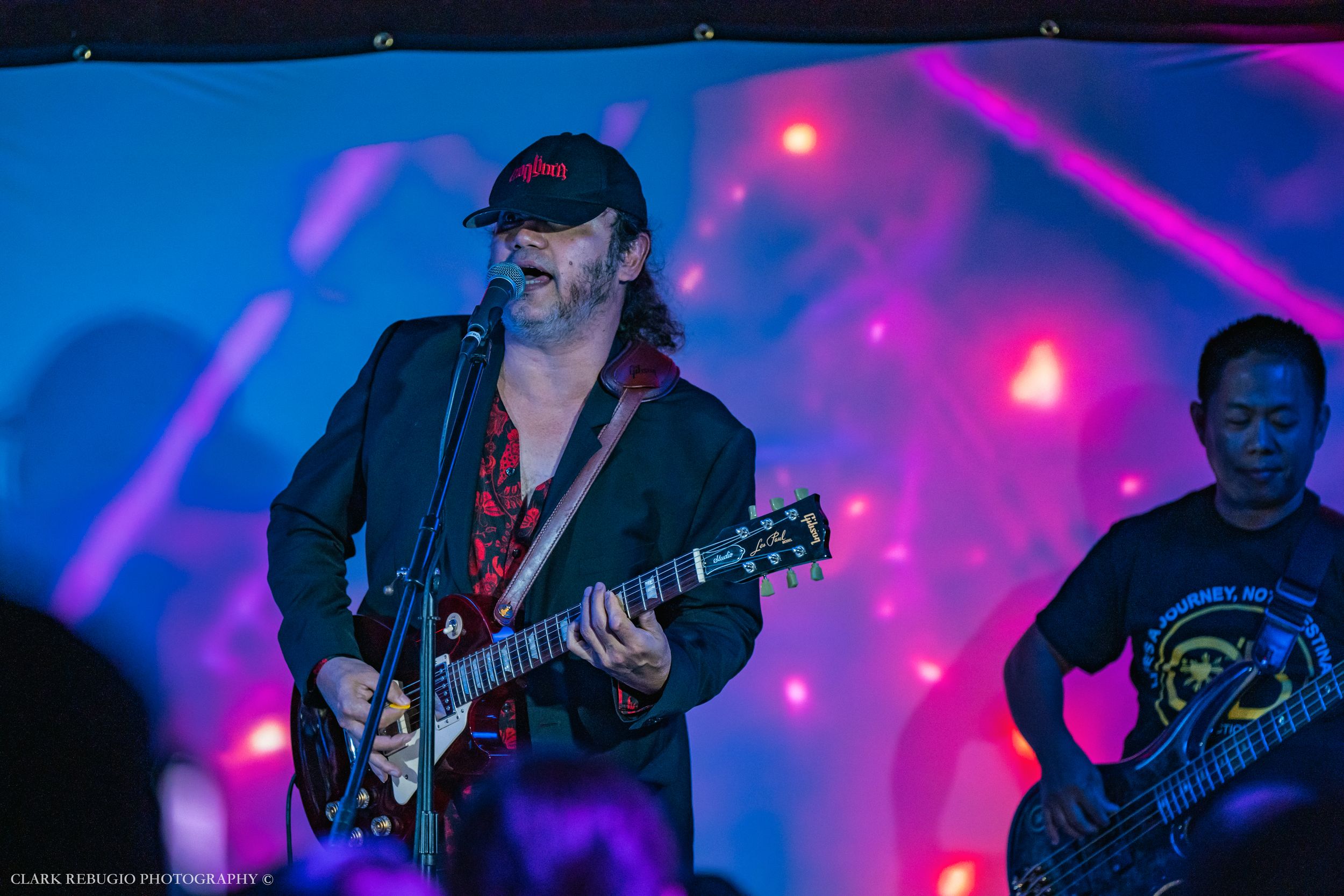 Male musician wearing a cap playing electric guitar and singing on stage under purple and blue lighting at a live concert.