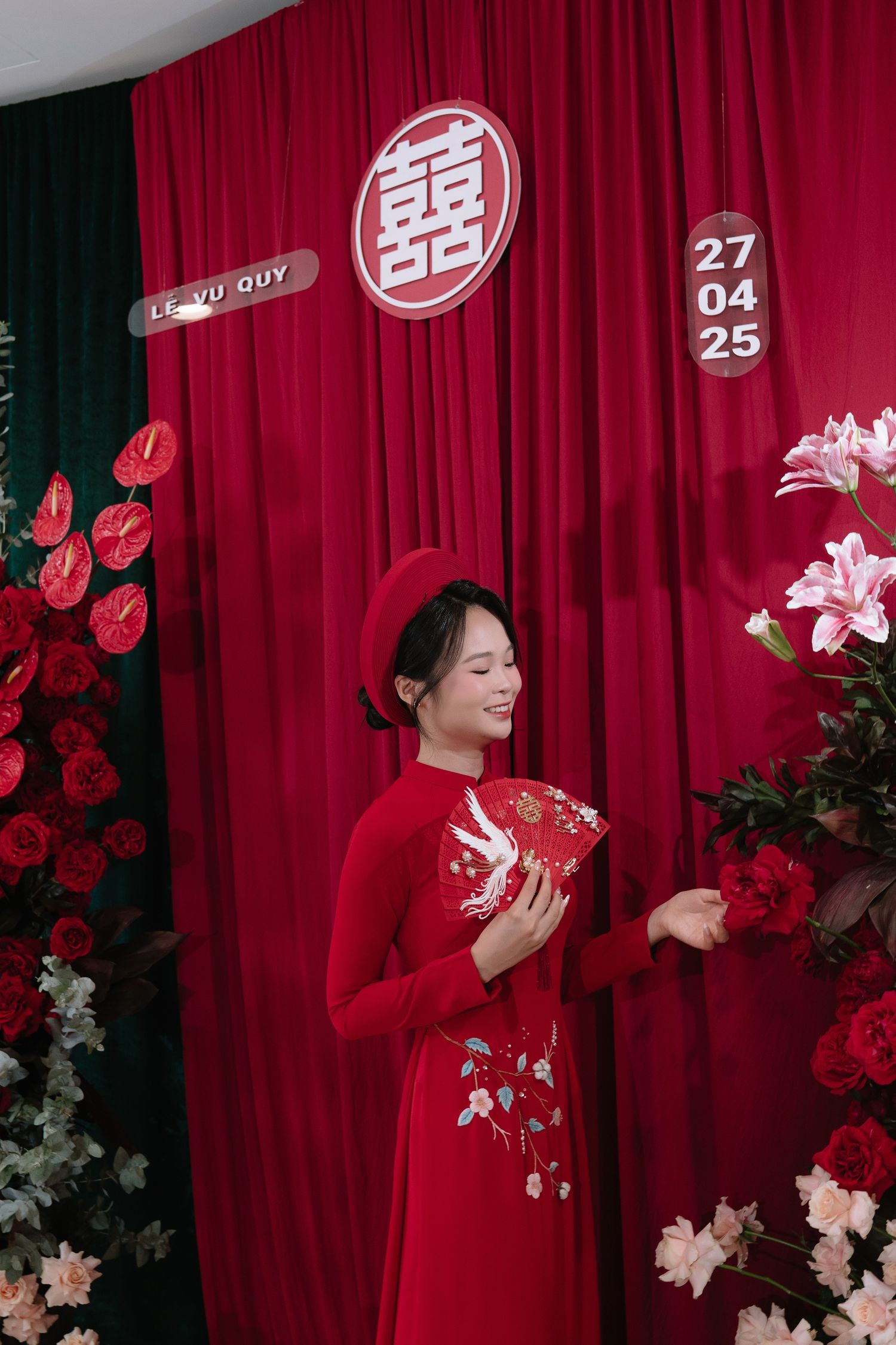Vietnamese bride in red ao dai smiles holding decorative fan at wedding ceremony backdrop.