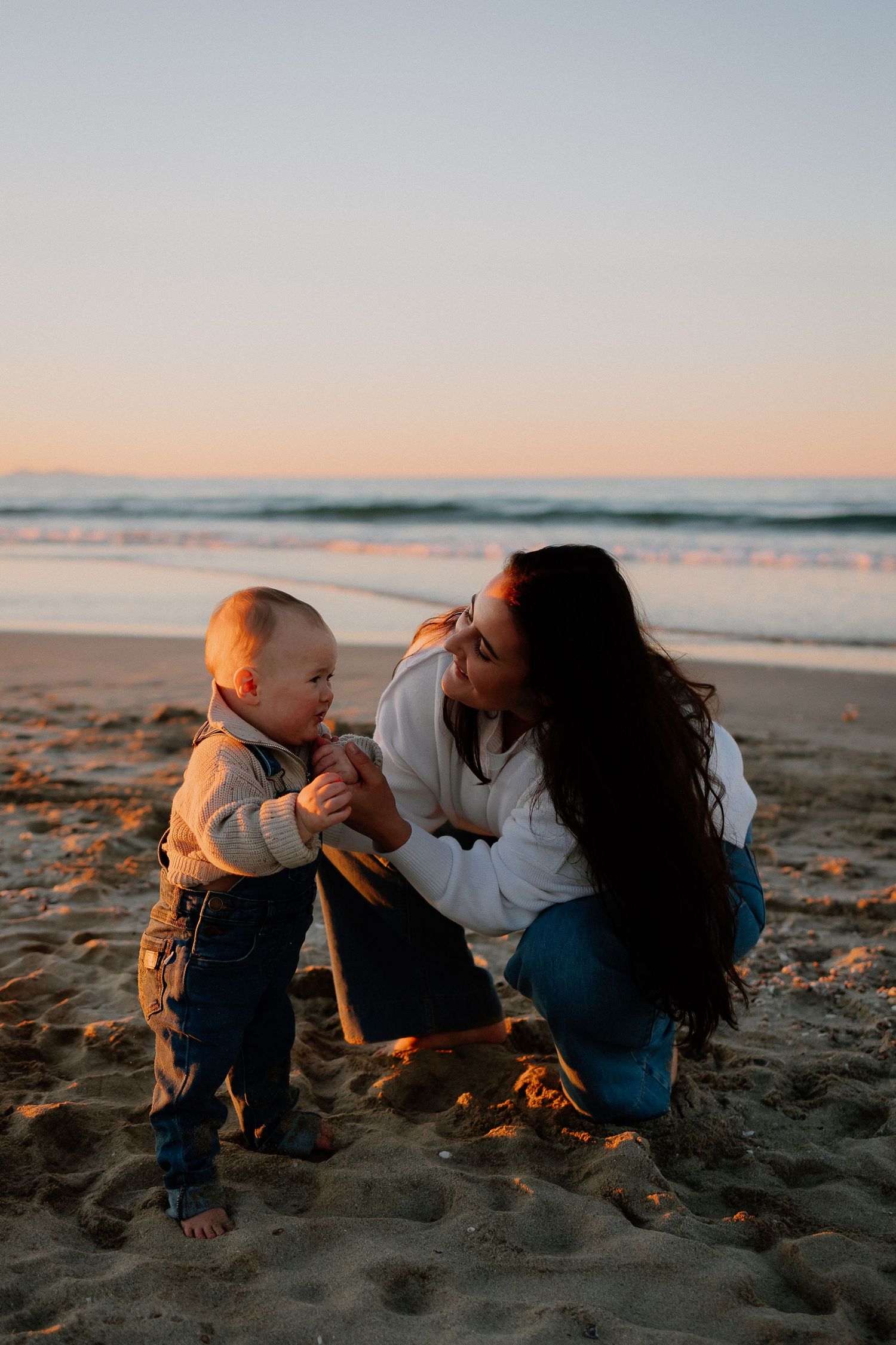 A tender beach moment at sunset with a toddler and adult sitting together on golden sand by the ocean.