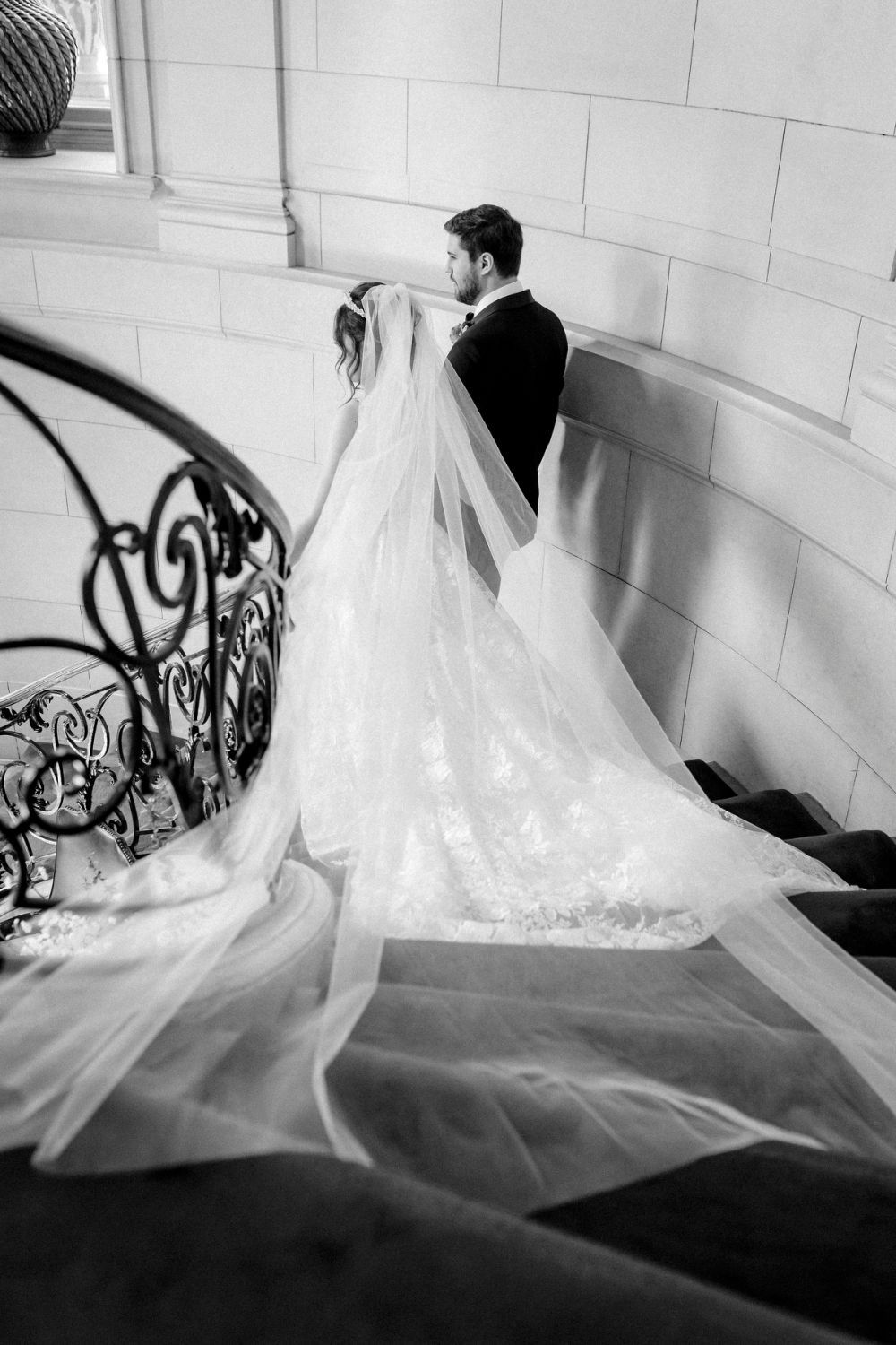 Black and white wedding photograph capturing a bride and groom ascending elegant stairs, with flowing veil trailing behind.